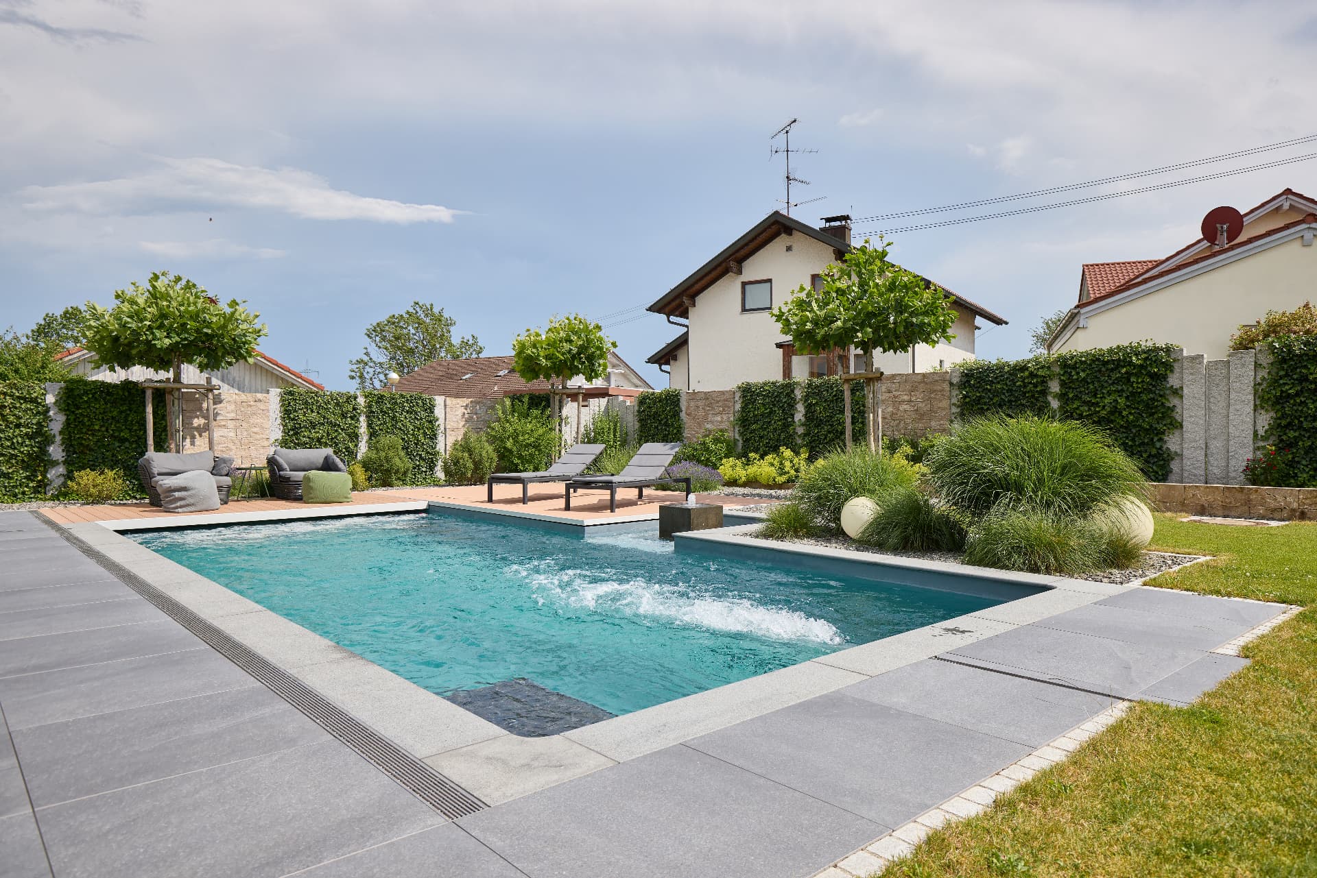 Pool mit grauer Plattenumrandung, Holzdeck und Bepflanzung im Garten